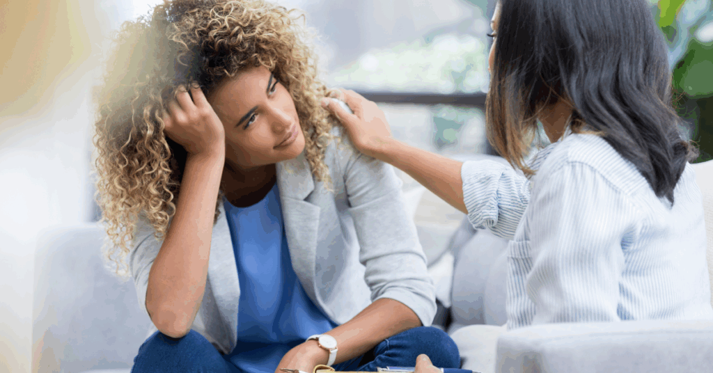 Brightly lit room with stressed looking woman and another woman's hand on her shoulder for comfort.