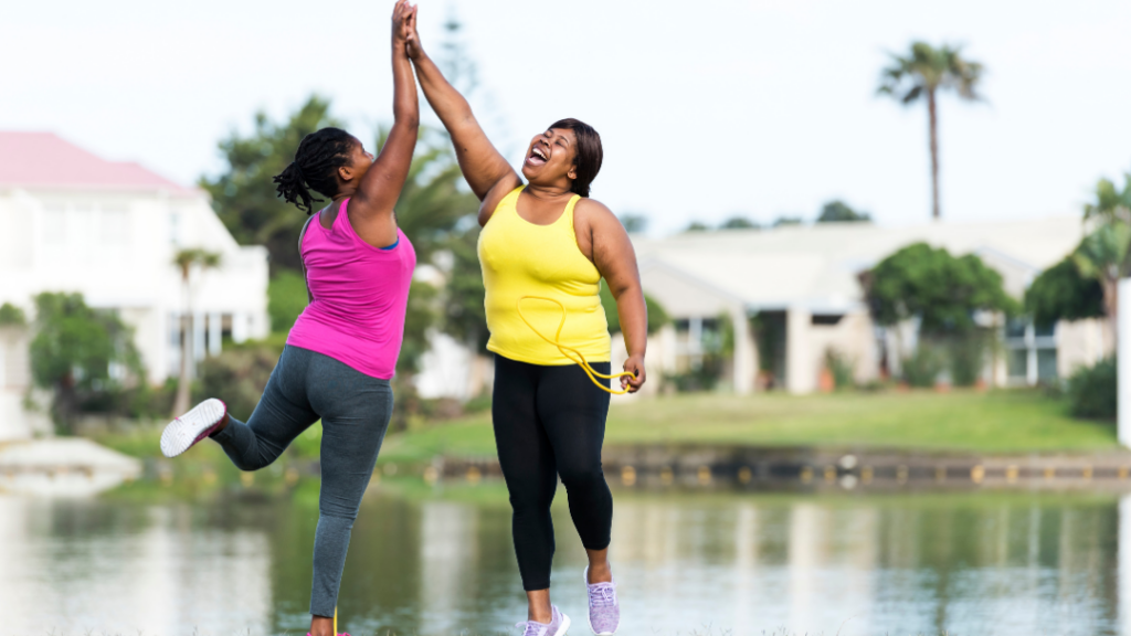 Two women in colorful activewear high-five while jumping outdoors beside a pond, smiling tailwind of a workout session.