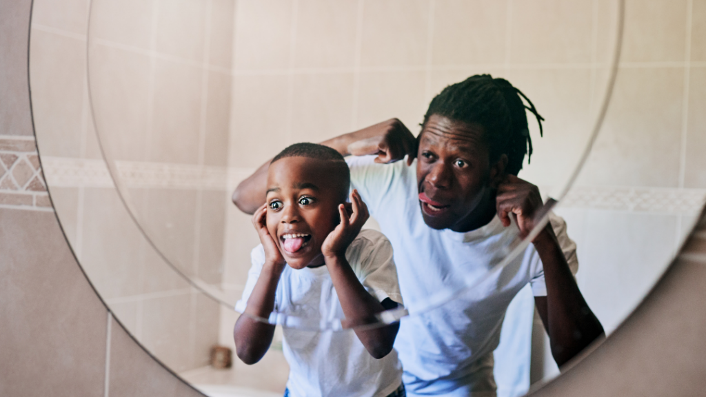 How to Support a Child’s Mental Health in Everyday Life - Father and son in a bathroom, leaning toward a round mirror and making silly faces at their reflection.