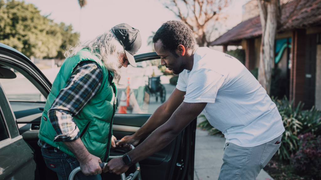 Life After a Stroke: The Emotional and Mental Health Side of Recovery - A caregiver helps an older man in a green vest and cap into a car, assisting with a walker near the open driver's door.