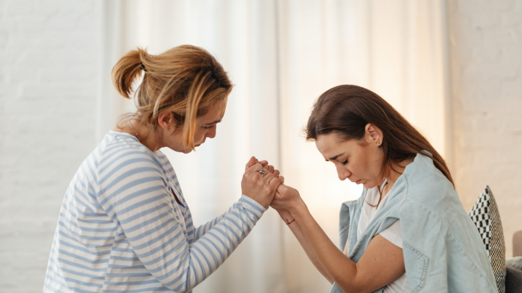 Supporting a Loved One Through Major Health Changes - Two women sit facing each other, gripping hands in a comforting, supportive moment in a bright living room.