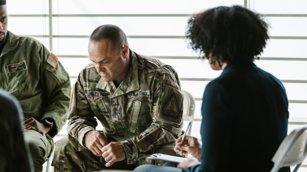 PTSD Is More Than Flashbacks: Understanding the Many Faces of Trauma - Soldier in camouflage uniform sits with hands clasped, looking down, as a civilian interviewer takes notes at a bright, windowed room.