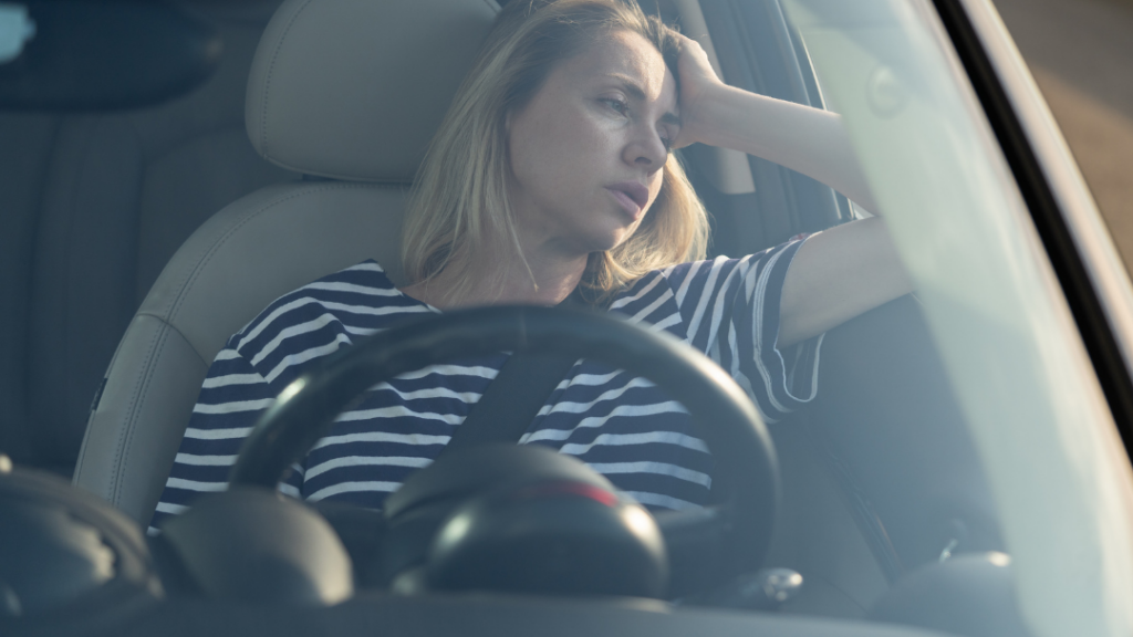Woman in a striped shirt rests her head on her hand in the driver's seat, looking exhausted inside a car.