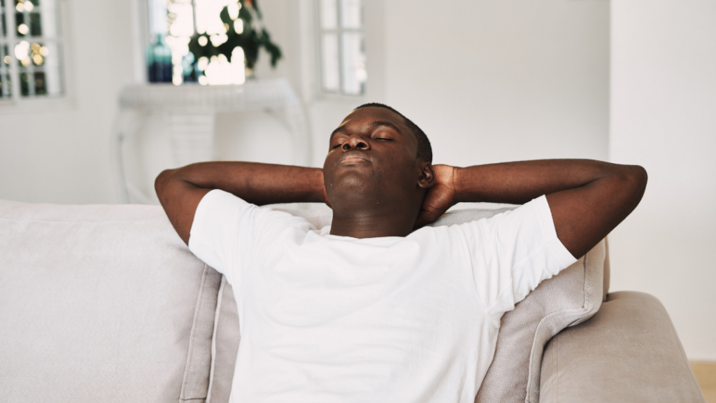 Man with white T-shirt reclining on a light sofa, arms behind head, eyes closed, relaxing at home