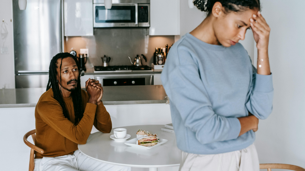 How Trauma Can Affect Relationships and Connection - Man seated at a white kitchen table with clasped hands, looking worried as a woman in a blue sweater stands nearby and holds her head; sandwich and coffee on the table behind them in a modern kitchen.