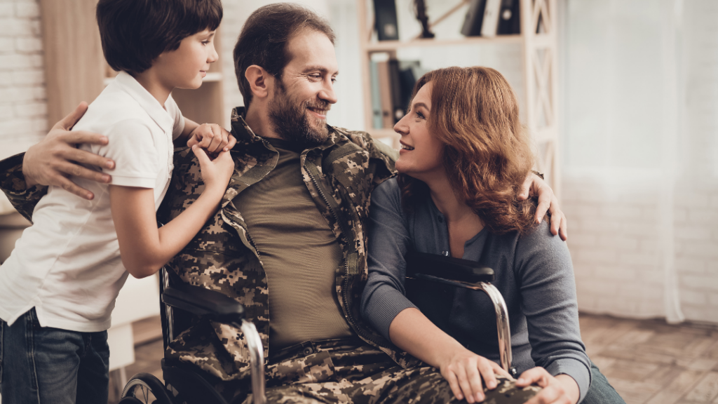 Smiling family: a man in a wheelchair wearing camouflage sits between his partner and their young son at home, all hugging the moment.