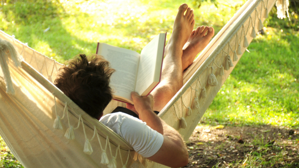 Man relaxing in a hammock outdoors, reading an open book in a sunlit garden.
