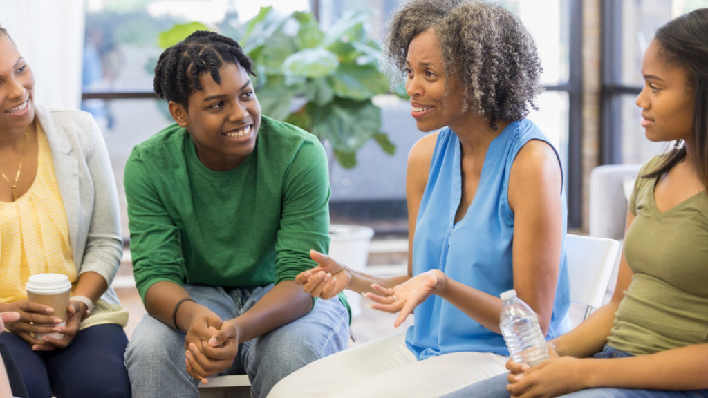 A diverse group of four adults sits in a circle, chatting and smiling in a bright room with a plant in the background.
