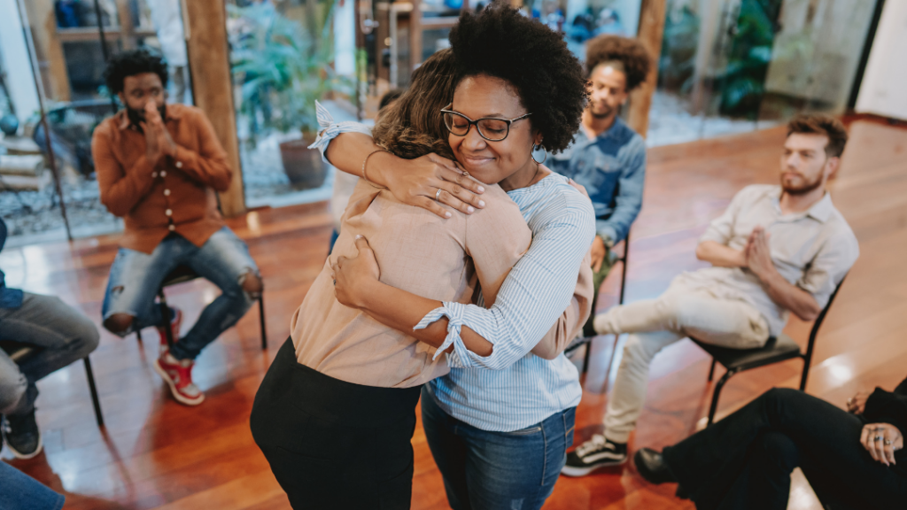 Two women share a warm hug as a circle of participants watches in a bright, open room.