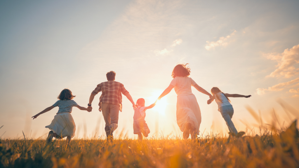 Family of five holding hands and running through a sunlit field at sunset.