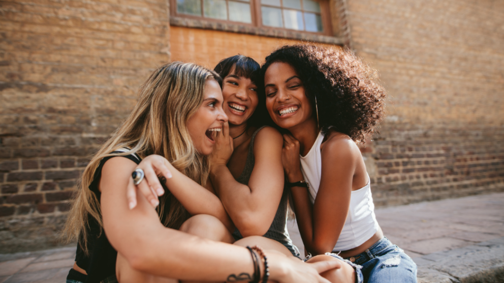 Three friends laughing together outdoors against a brick wall.