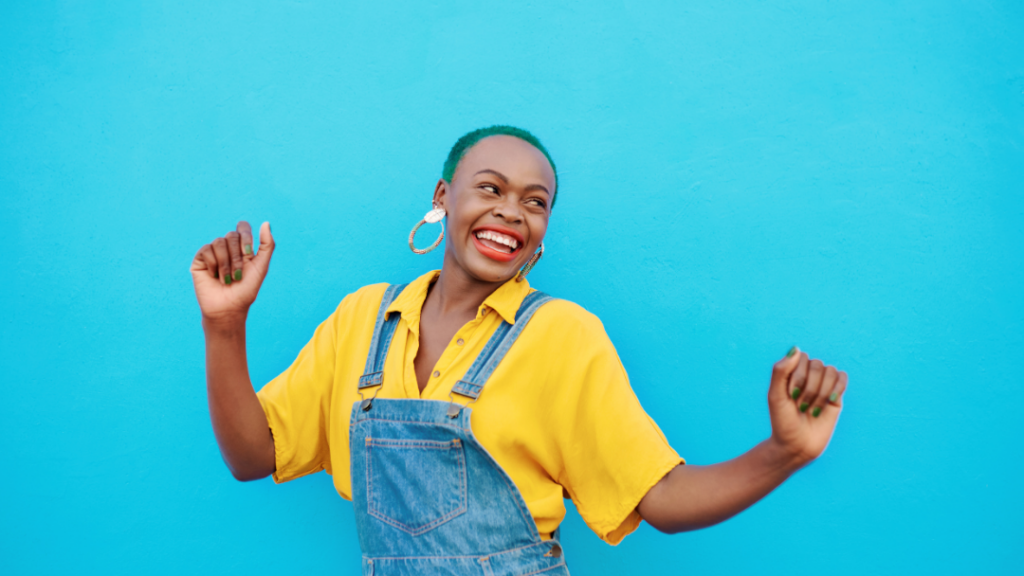 Joyful woman with green hair, wearing a yellow shirt and denim overalls, dancing and smiling against a bright blue wall with large hoop earrings and green nails.