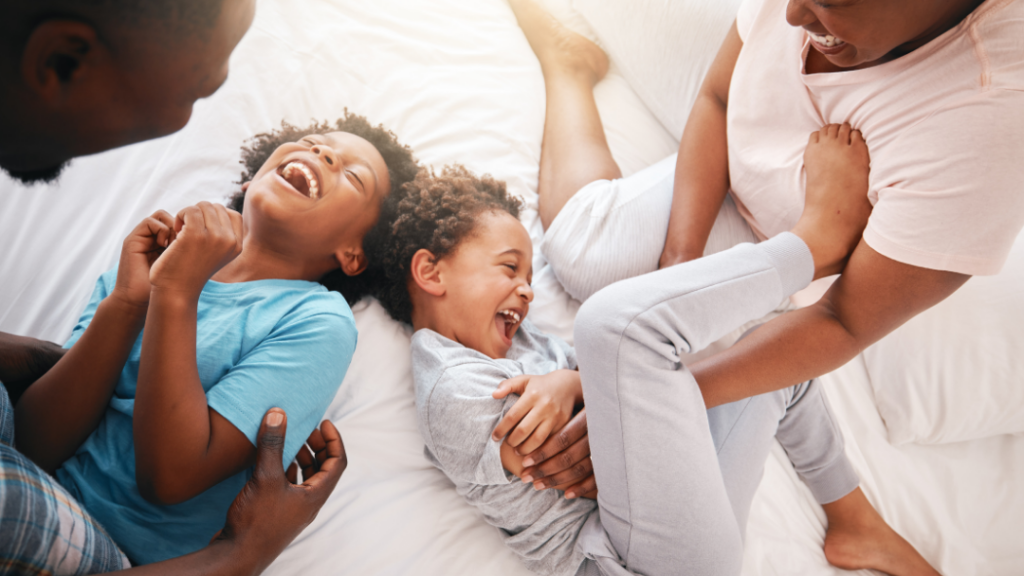 A joyful family lying on a white bed, laughing together as a parent plays with the two children.