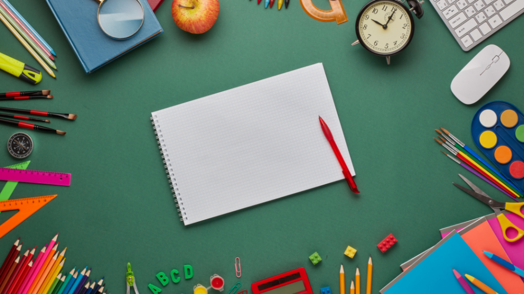 Open spiral notebook with a red pen on a green desk surrounded by school supplies like pencils, rulers, paints, and scissors on a flat lay.