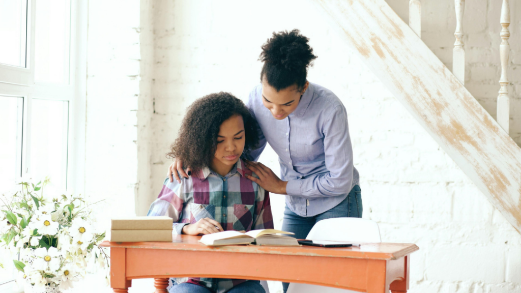 Two young women studying together at an orange desk by a bright window; one stands beside the seated partner, offering guidance.