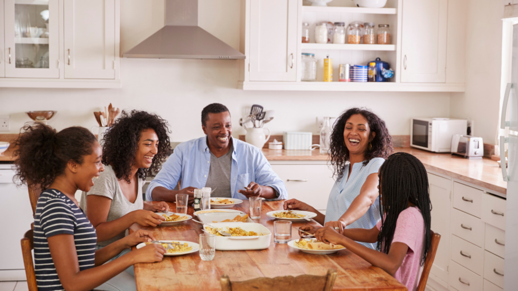 A diverse group of friends/family laughing and sharing a meal at a sunny kitchen table.