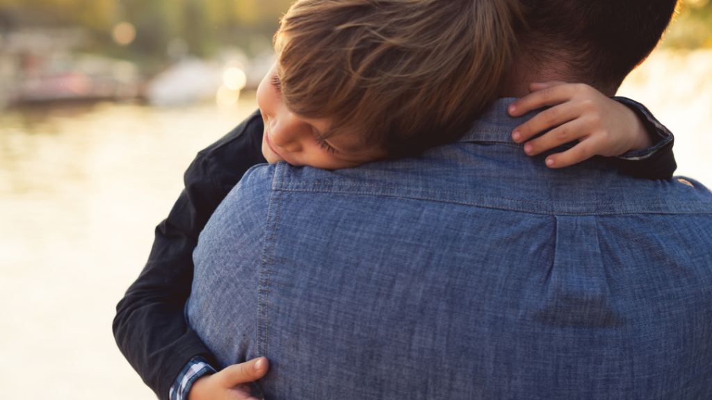 Child hugs an adult from behind by a sunlit lake at golden hour with a denim shirt visible on the adult.