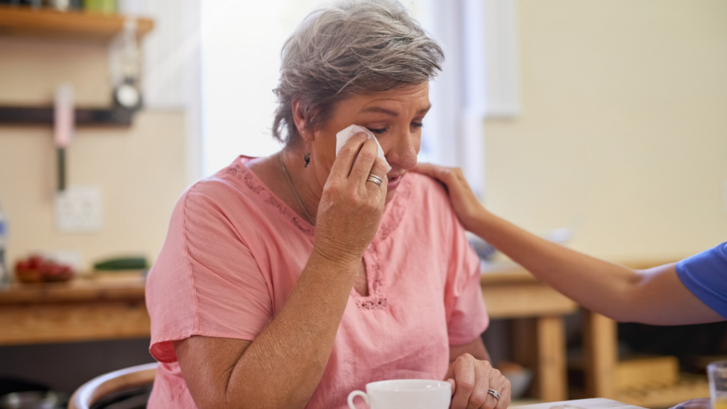Elderly woman in a pink blouse wipes her eye with a tissue while a caregiver in blue places a reassuring hand on her shoulder