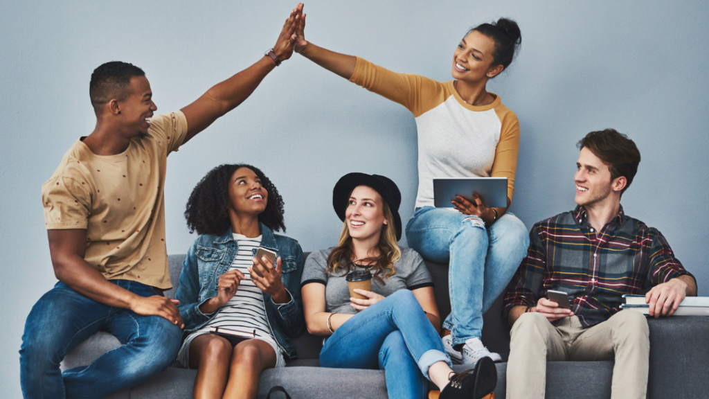 Five diverse friends sit on a gray sofa two in the center high five while others smile and chat with devices in hand