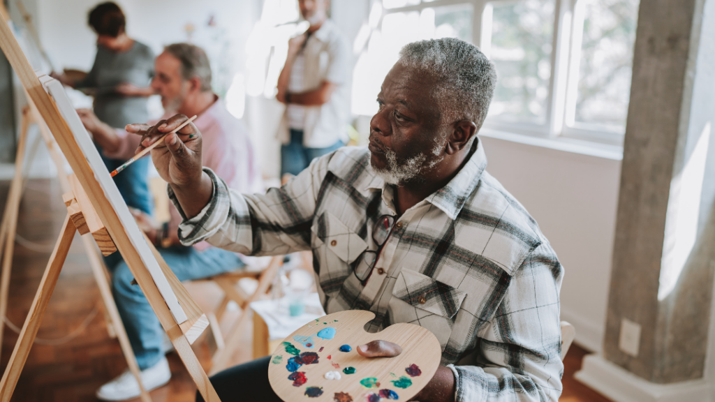 Life After a Crisis Rebuilding Emotional WellBeing Older man with a gray beard painting at an easel in a bright group art class others working in the background