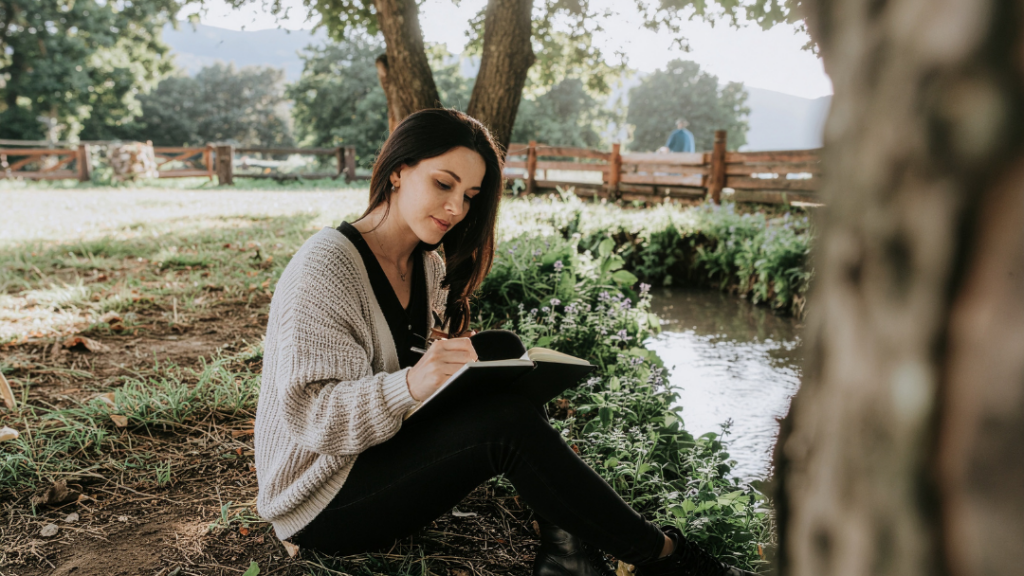 Mental Fitness Building Emotional Resilience Every Day Woman sitting on grass by a pond writing in a notebook while wearing a beige cardigan