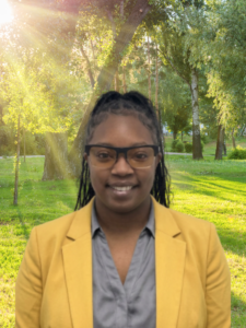 Headshots for Website Smiling person in a yellow blazer and glasses stands in a sunlit park with trees in the background