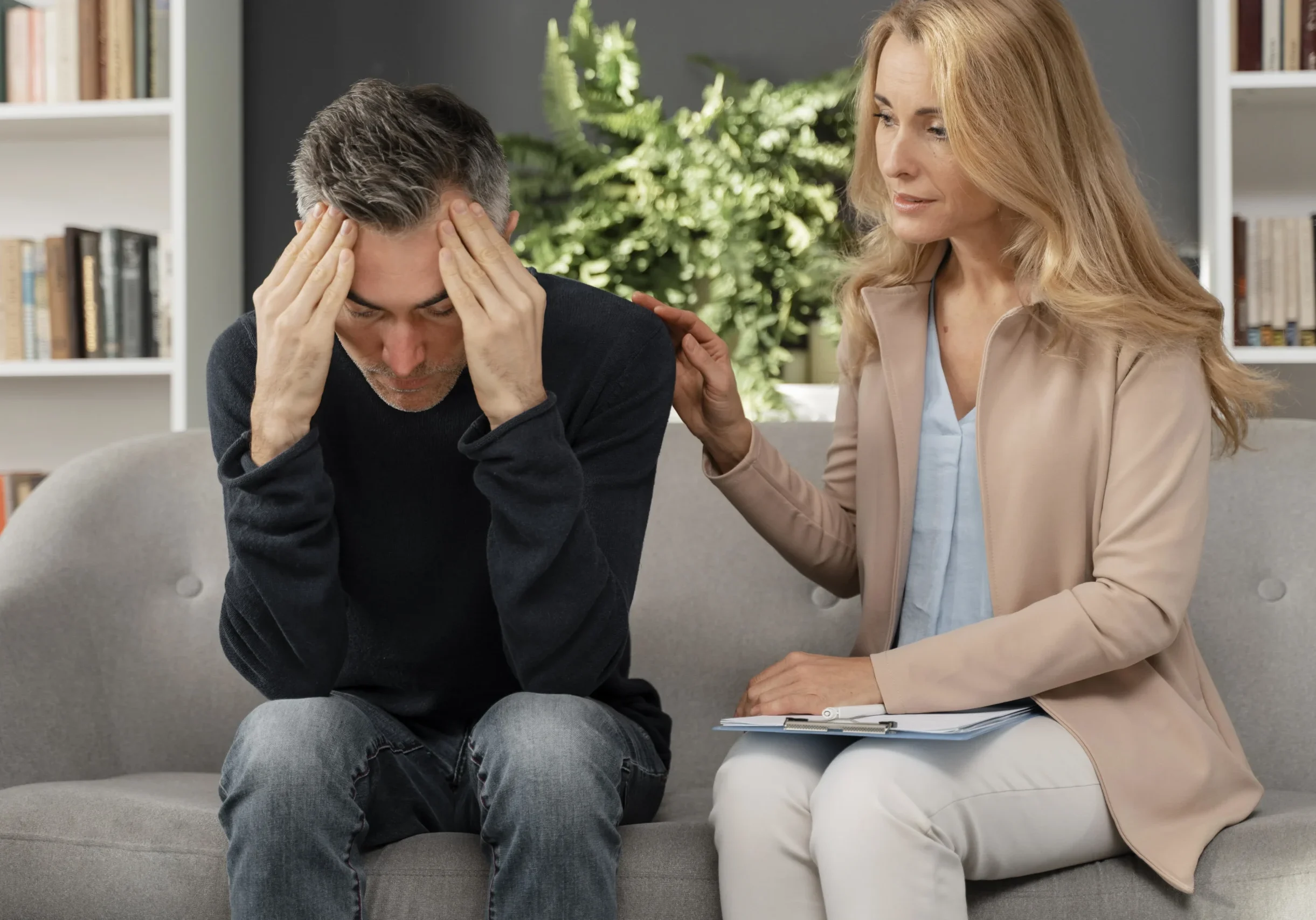 distressed man sitting on a couch with his head in his hands while a woman therapist offers comfort with a hand on his shoulder and a notebook nearby in a living room setting